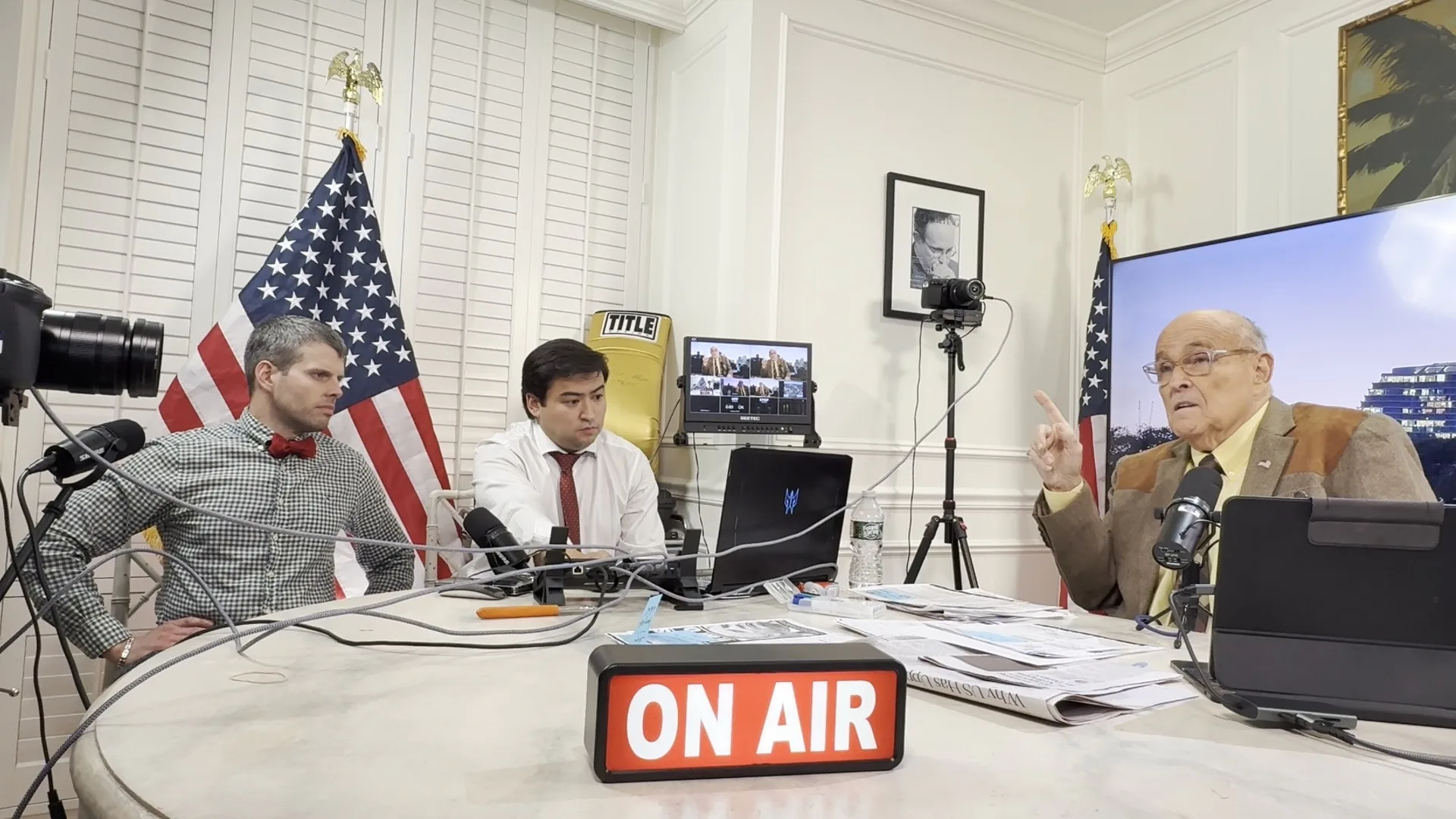 Stephen Schumacher and Ted Goodman listen as Rudy Giuliani speaks at a microphone during America’s Mayor Live