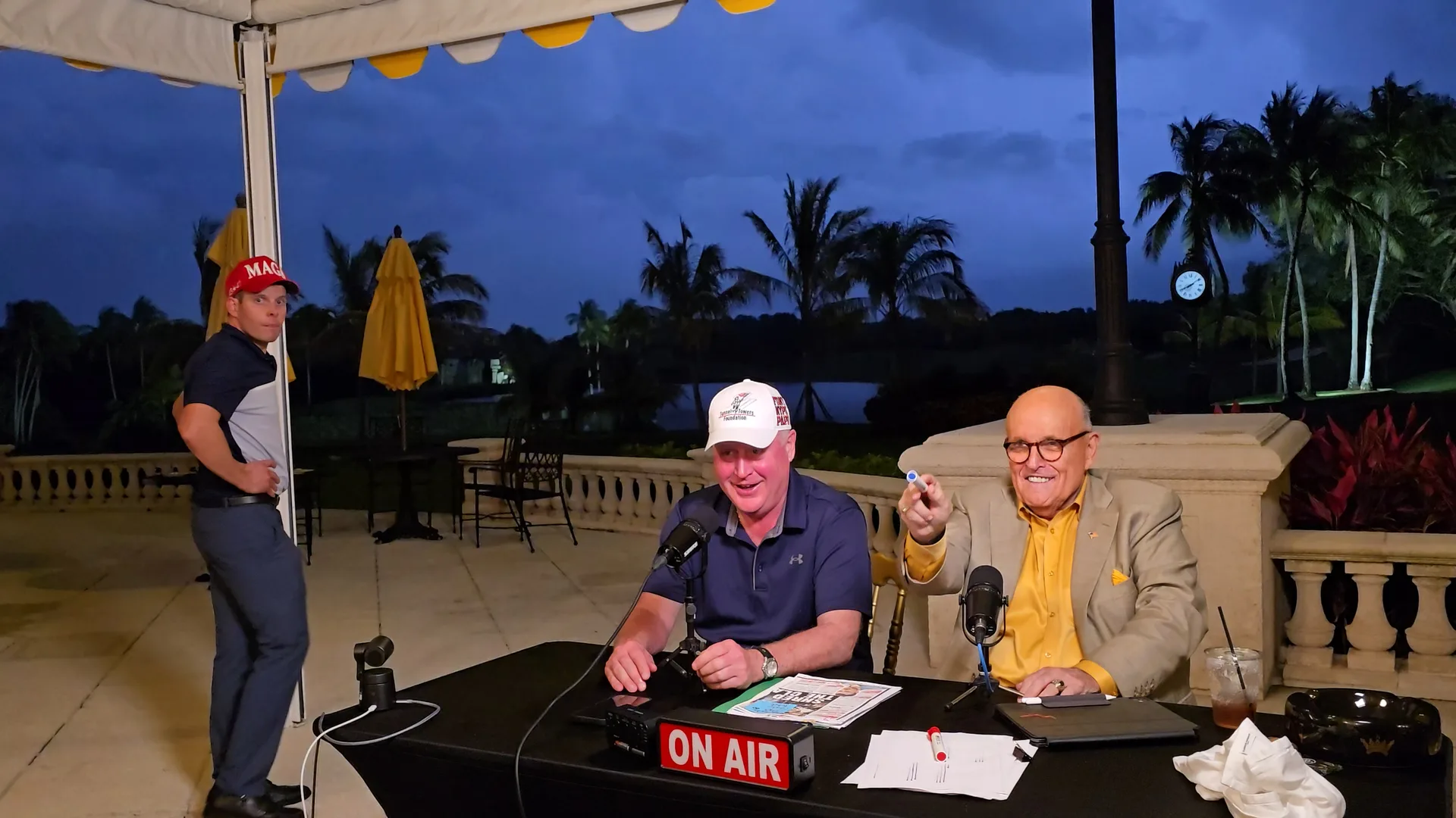 Rudy Giuliani and a guest seated at outdoor microphones while Stephen Schumacher stands beside the America’s Mayor Live production setup at Trump International