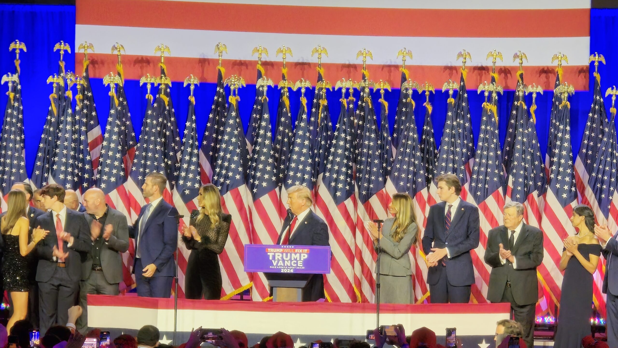 A national campaign victory stage surrounded by American flags and Trump campaign signage